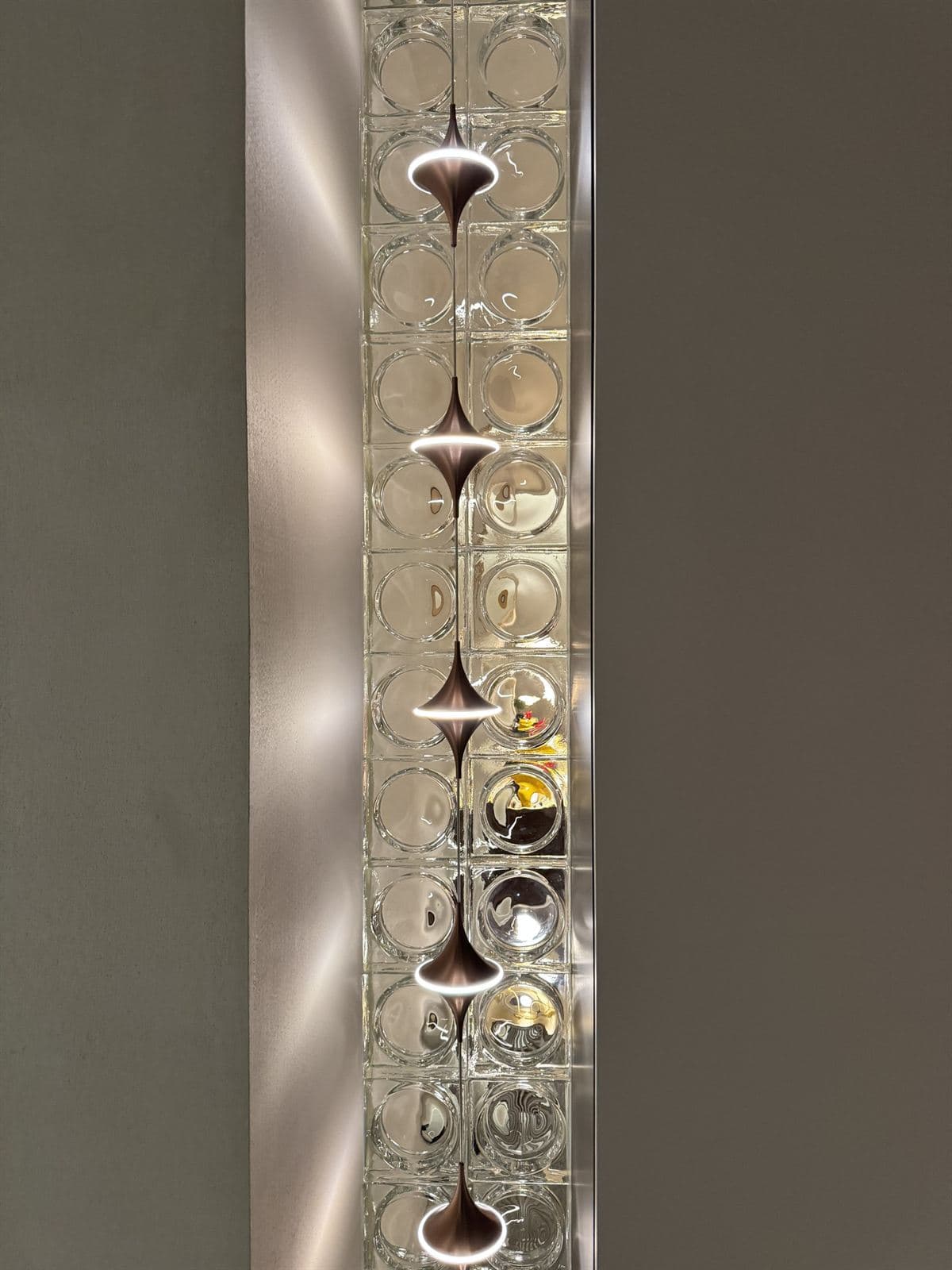 A vertical arrangement of glass blocks lit from within by three modern metallic GUOCIO pendant lights