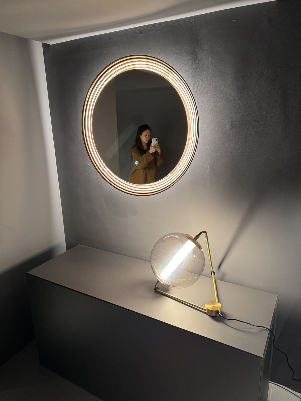 A round, back-lit GUOCIO Halo art mirror with a ribbed brass frame on a charcoal wall, above a brass table lamp with a clear glass globe