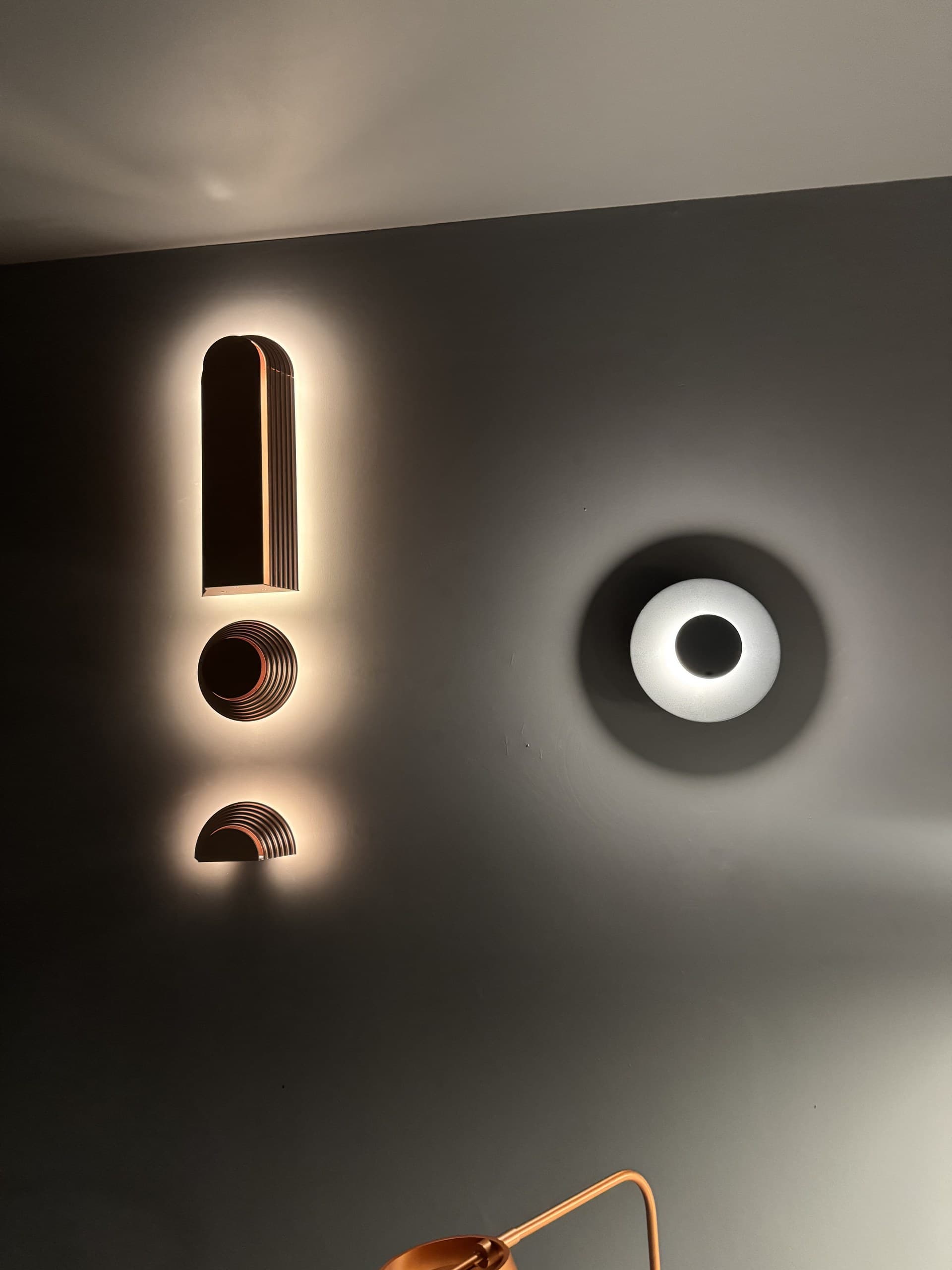 Two distinct wall-mounted light fixtures illuminate a dark gray wall, one shaped like an exclamation mark and the other a circular globe.