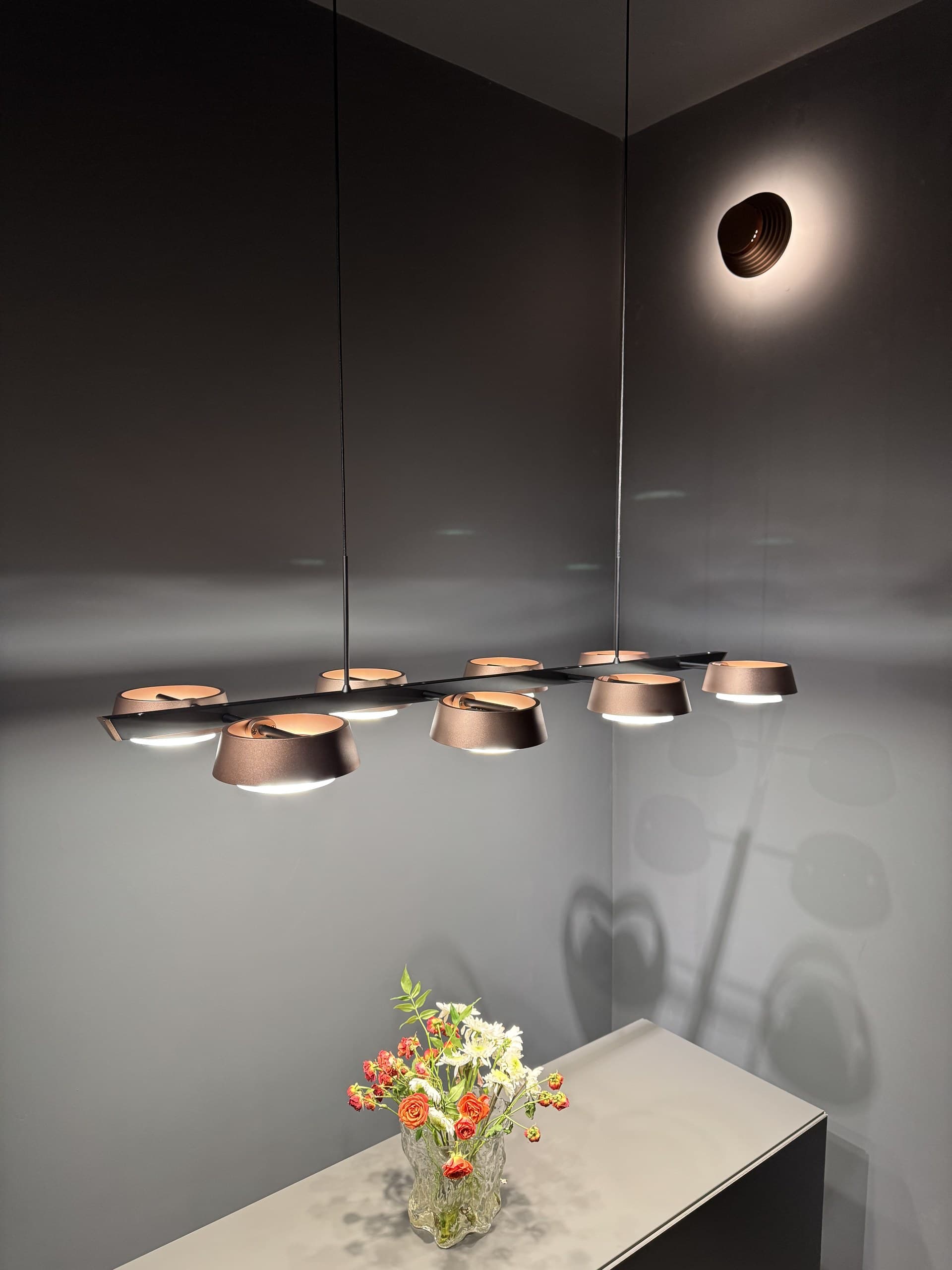 A linear pendant light with multiple bronze-colored shades illuminates a corner of a room with a vase of flowers on a counter.