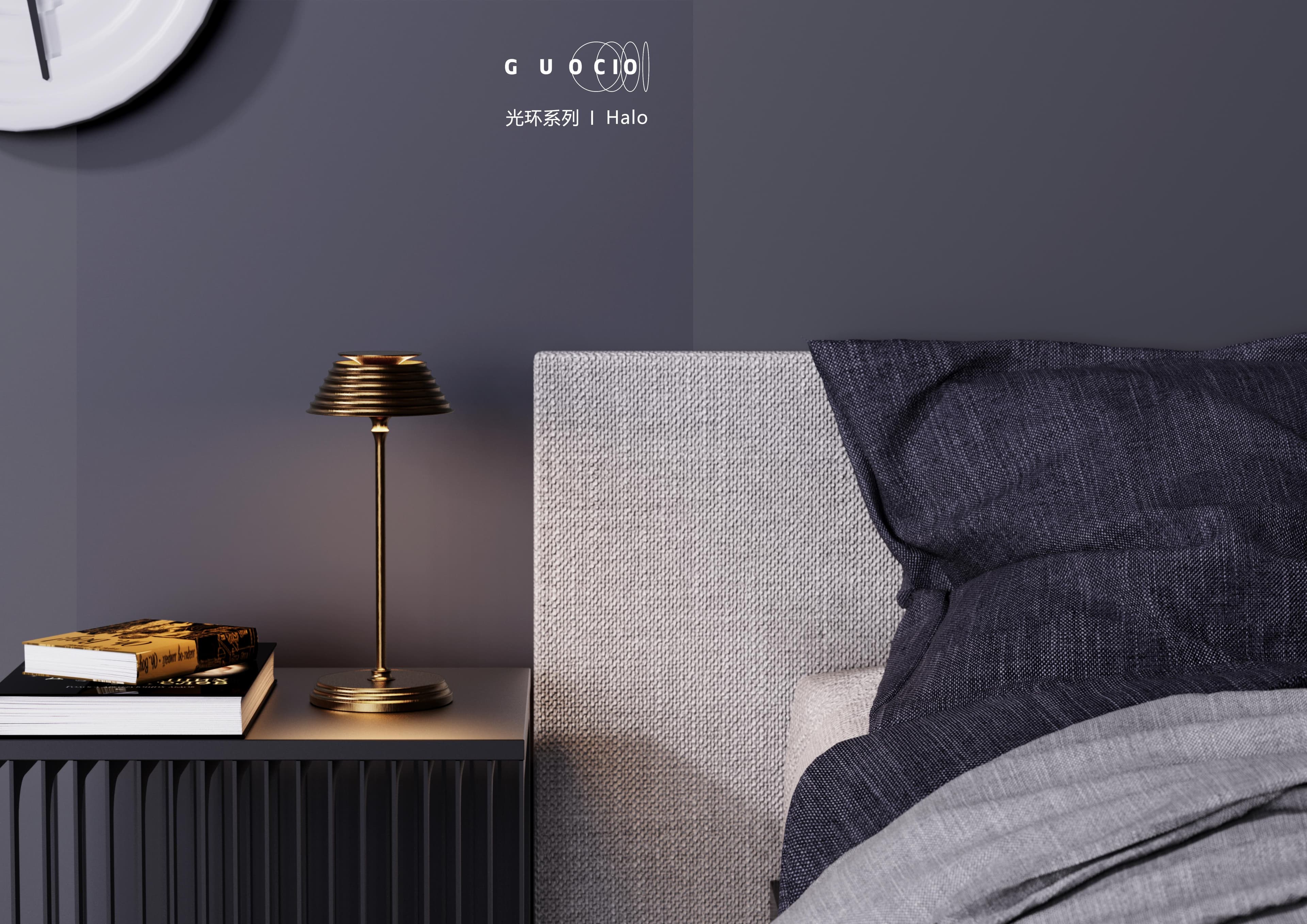 A bedside table with a golden lamp and two books, next to a bed with a grey headboard and dark blue and light grey bedding.